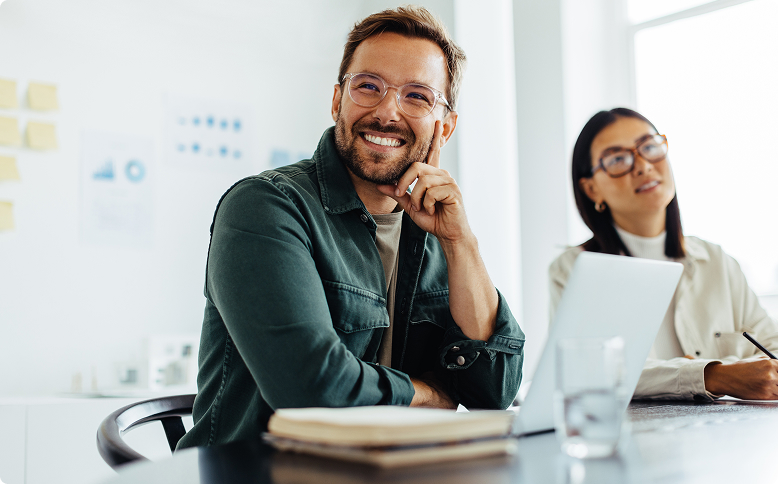 Büro: Lachender Mann mit Brille und Bart in grünem Hemd, sitzt am Besprechungstisch, rechts von ihm eine Frau.